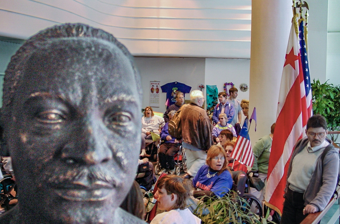 The bust of Martin Luther King is in the foreground with colorful people crowded behind. 