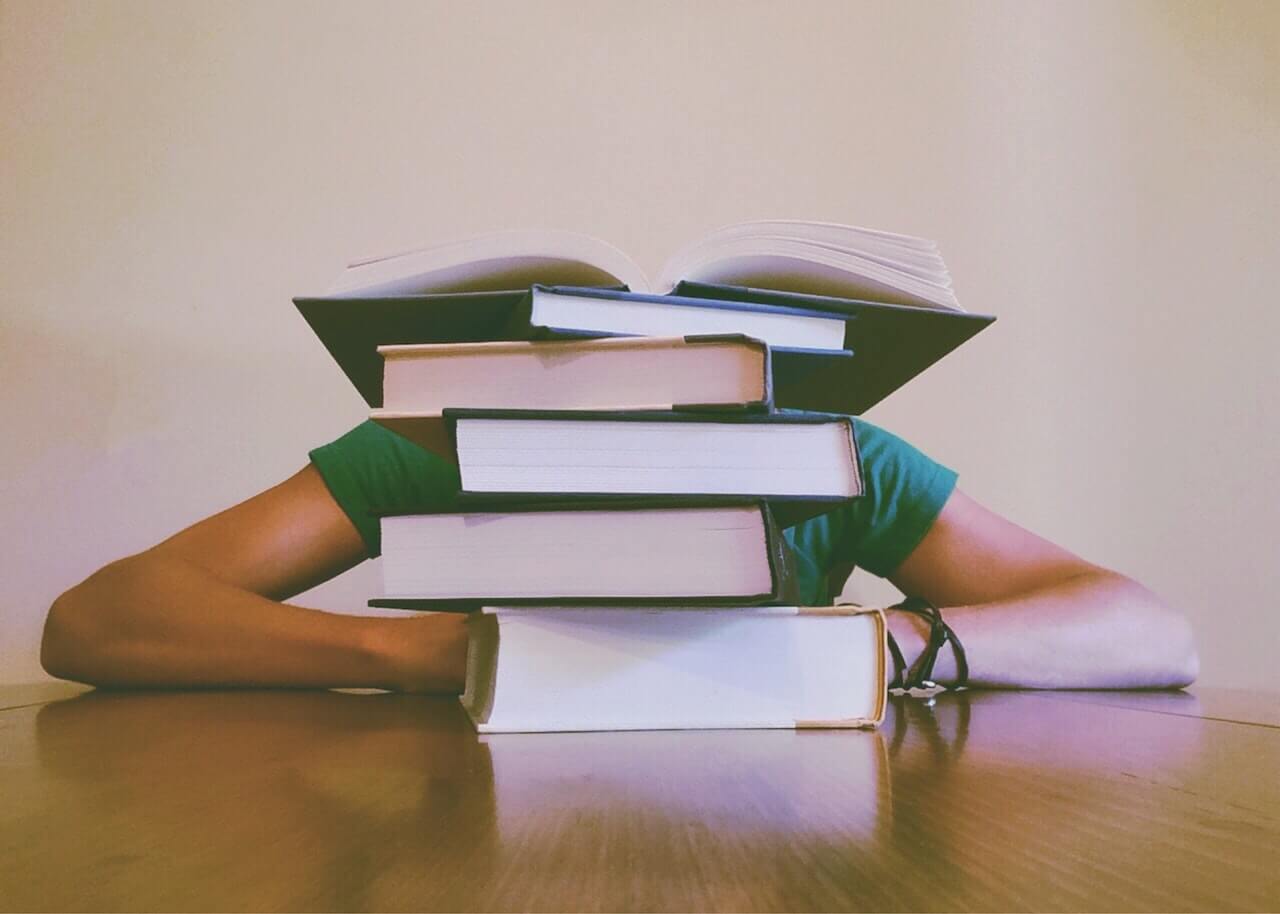 A student with arms spread on table is hidden behind a stack of several books, with the top one open.