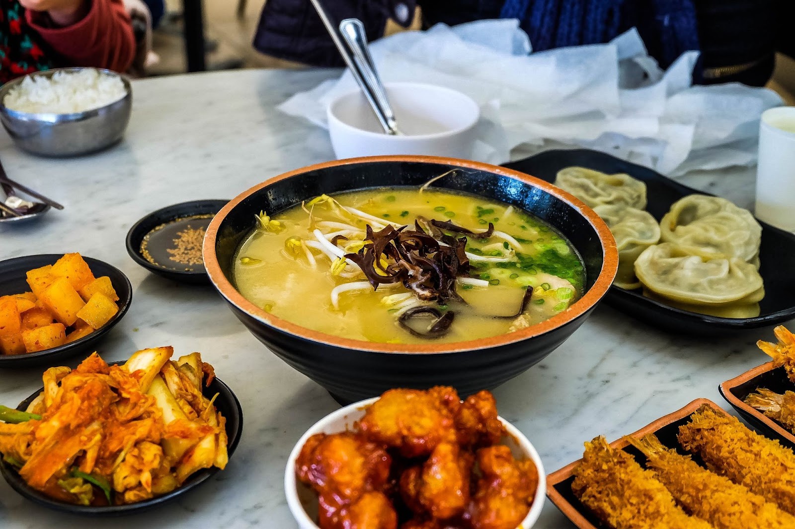 Korean cuisine on a marble countertop, including soup, dumplings, chicken and fried battered seafood
