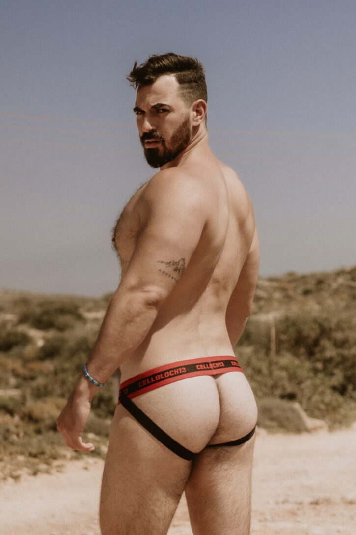 Thick Macho wearing a black and red jockstrap from cellblock 13 on the beach while posing and looking over his shoulder at the camera