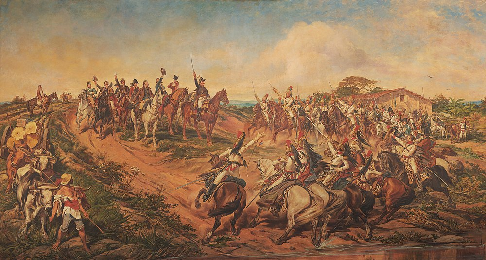 Dom Pedro I ergue sua espada acompanhado de outras pessoas montadas em cavalos ao seu redor em um campo.
