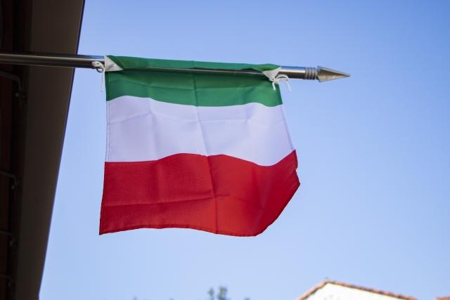 イタリア国旗が鉄のやりに結ばれ掲げられた写真。背景の空が青く象徴的。