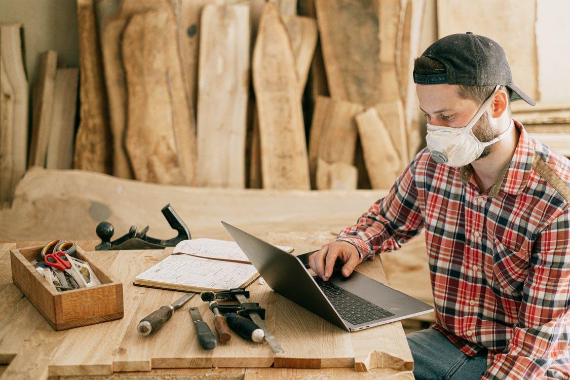 A carpenter in a white mask sitting at his laptop surrounded by tools and wood.