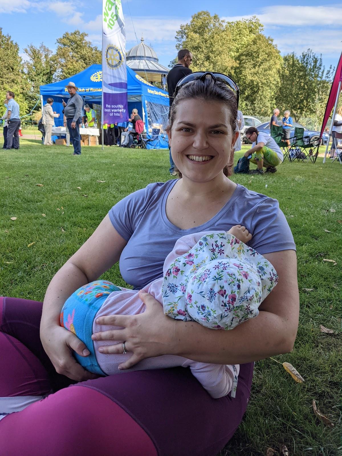 Fiona feeding her daughter after finishing a charity walk. Fiona is in exercise leggings and a purple t-shirt. Her daughter is wearing a flowery, floppy sun hat, bright blue  flowery leggings and a pink top.