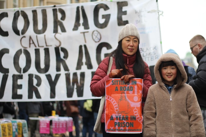 A mother and son smile beside a banner that reads Courage calls to Courage Everywhere. The Mother holds a sign that reads: Sitting in a road. A year in prison.