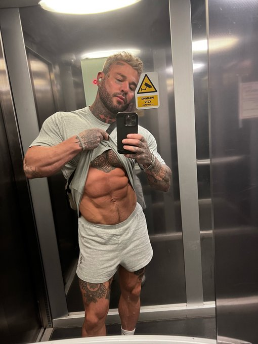 Danny Starr taking a mirror selfie in a bathroom wearing grey sweat shorts and lifting his shirt to reveal his abs