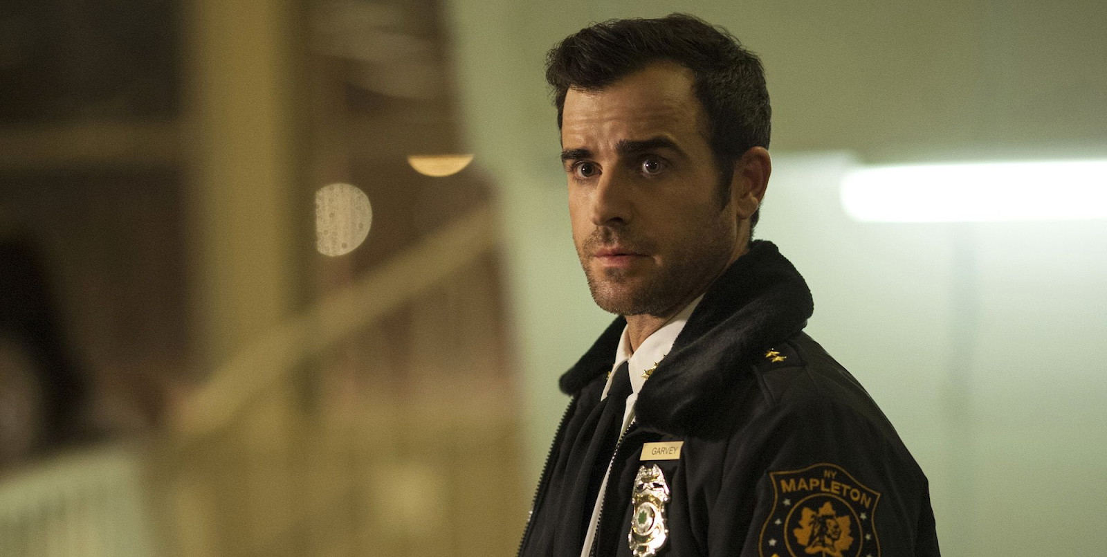 A screen still from The Leftovers, featuring Kevin Garvey, played by Justin Theroux, in his police uniform. He stares past the camera and the background is blurry and out of focus.