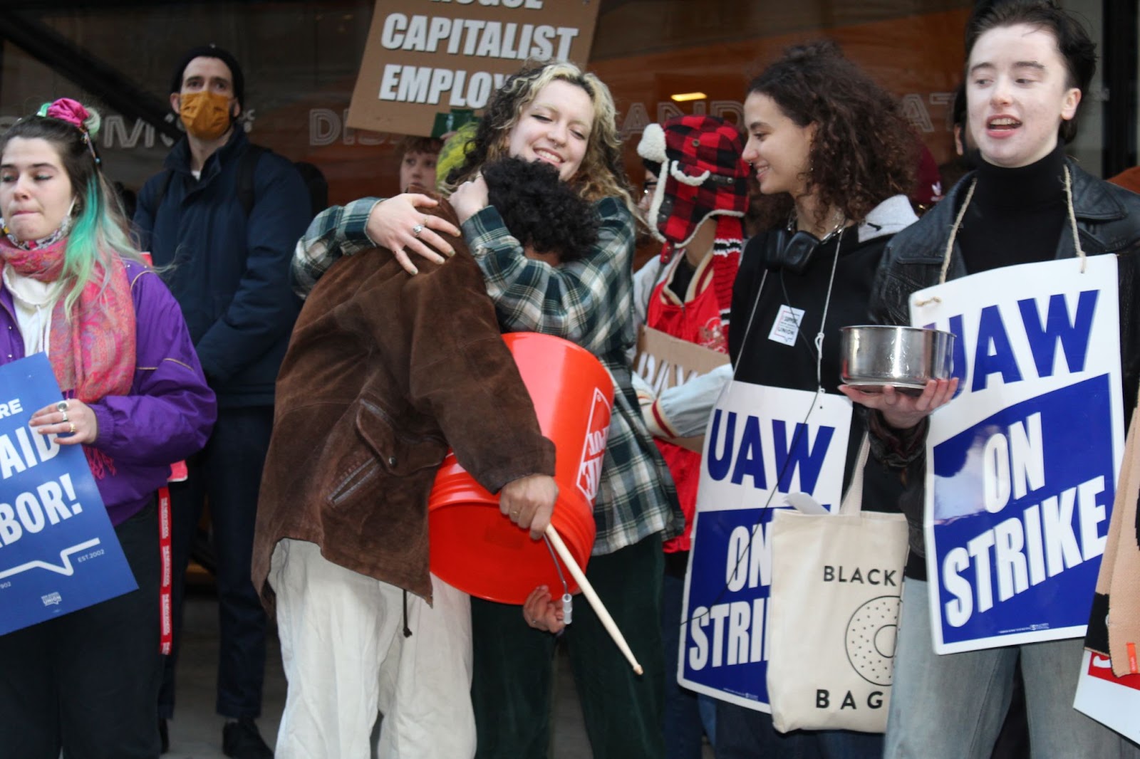 Students embrace at the part-time faculty strike. One holds a bucket as a drum, another holds a tin pot.