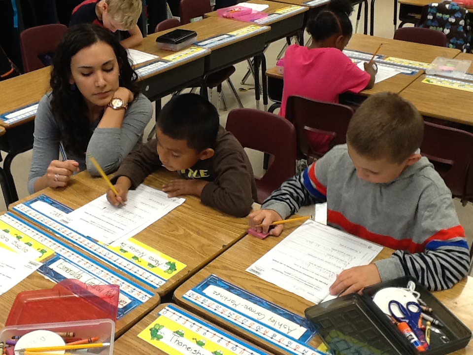 Elementary students work at their desks with a teacher helping