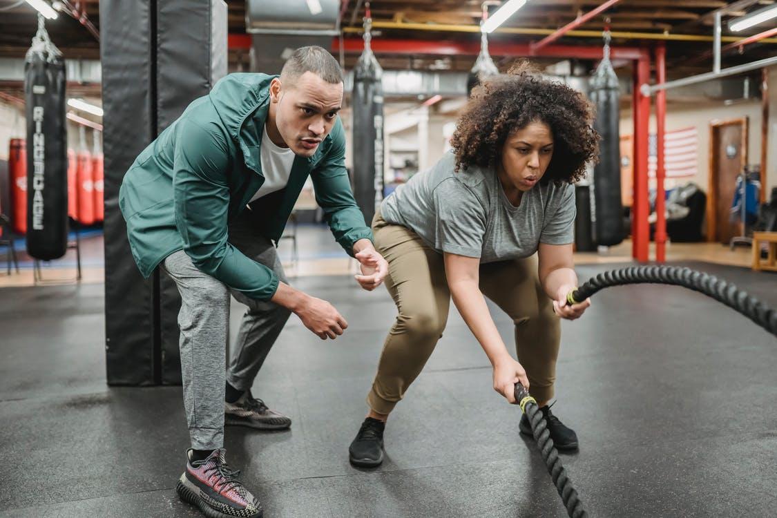 Determined black woman exercising with battle ropes near male coach