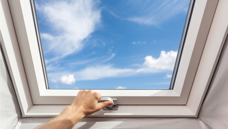 A man’s hand is securing an attic window that was recently installed during an attic renovation project. A bright blue sky with wispy white clouds is seen through the window.