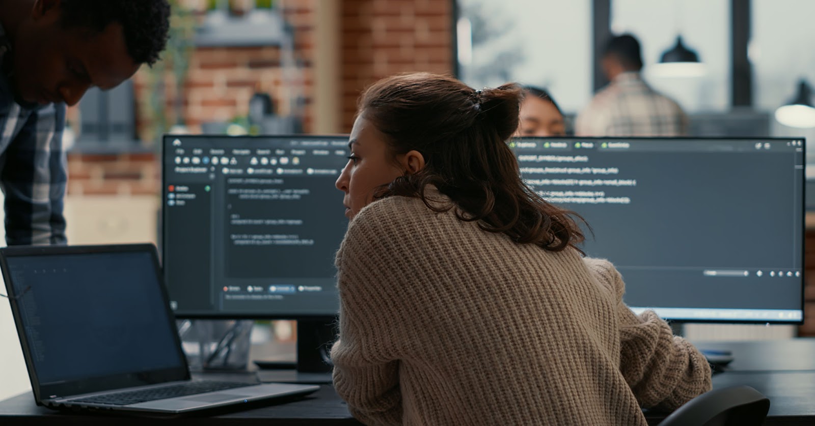 a woman in tech works hard on a line of code with double monitors and a laptop open
