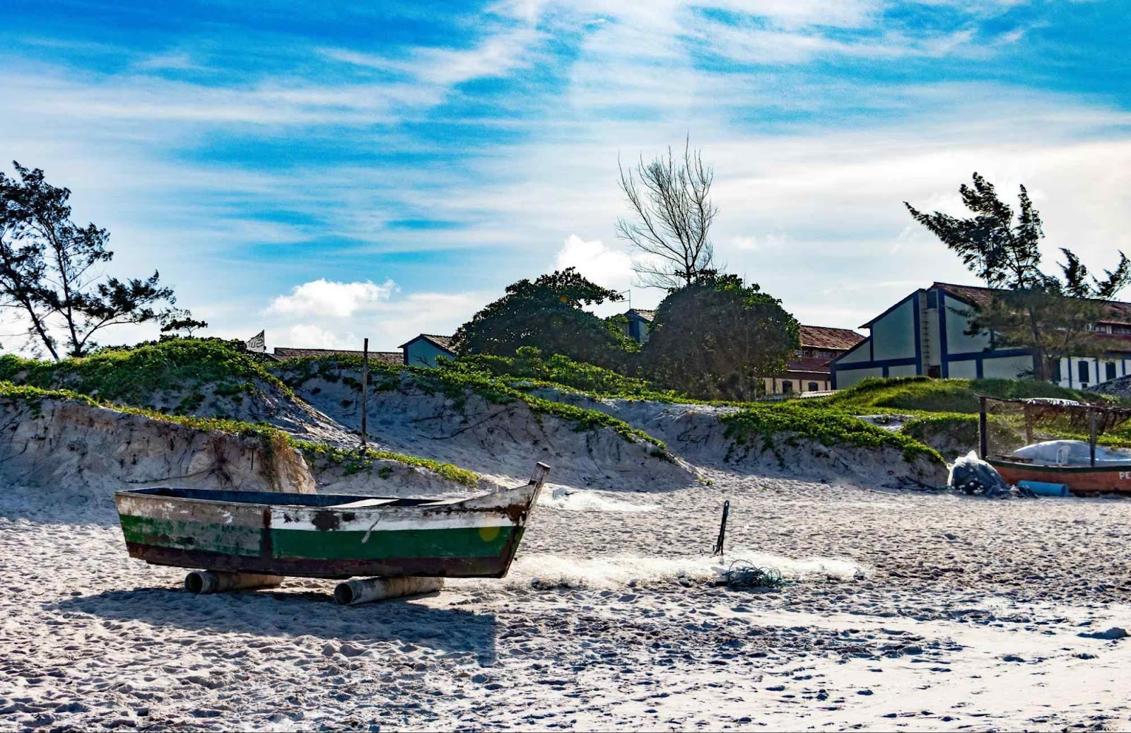 Pequeno barco colocado nas areias da Praia do Peró, em frente às pequenas dunas com vegetação em seu topo. Céu azul com nuvens brancas