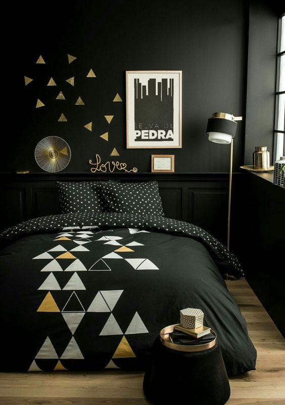 Quarto com cama de casal, paredes pretas, piso de madeira e acessórios preto e dourado.