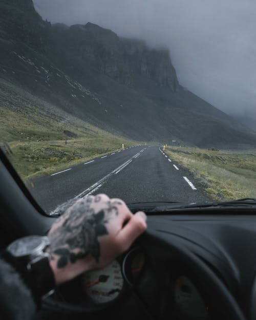 Crop anonymous traveler with tattooed hand on car steering wheel driving along road near mountains on overcast gloomy weather