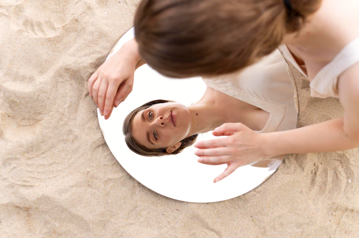 #DescriçãoDaImagem: fundo de areia clara com um espelho redondo em cima e uma mulher branca, de cabelos presos olha seu reflexo nele. Foto: Freepik.