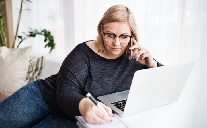 A woman researches Denver movers online while talking on the phone and taking notes.