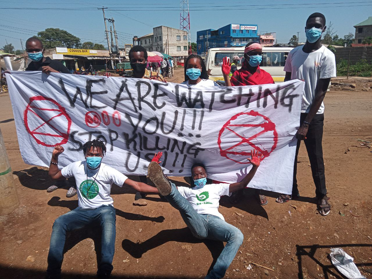 A group of XR Kisumu rebels hold a sign reading “We are watching you!!! Stop killing us!!!” Two rebels lie on the ground in front of the sign, one with his hands painted red