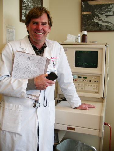 A male-presenting individual wearing a doctor's coat and holding a stethoscope and clipboard. He is leaning on a piece on medical equipment in what appears to be a doctor's office. 