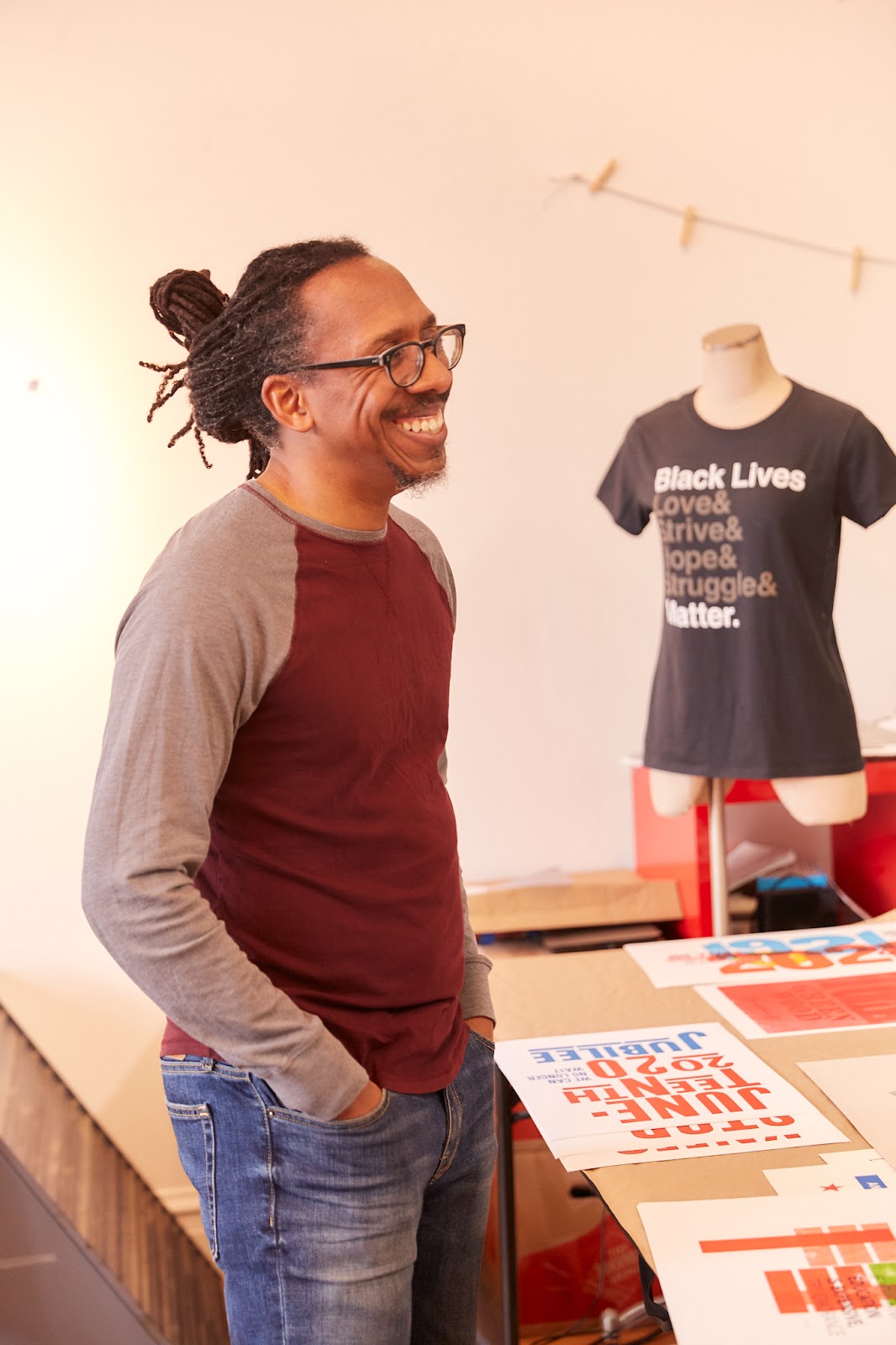 Image: Ben Blount smiling in his studio. He wears a long sleeved maroon and grey shirt and denim jeans, with his hands tucked in his pockets. To his right, is a table with a variety of test prints in mostly reds and blues. Above the table is a mannequin wearing a black t-shirt that says "Black Lives Love& Strive& Hope& Struggle& Matter." Photo by Sarah Joyce.