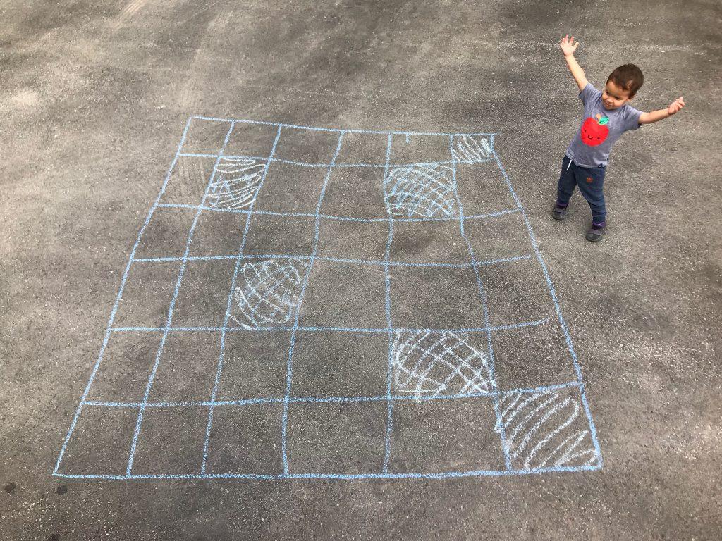 This shows a little boy beside a 6x6 grid of chalk squares on asphalt. Some of the squares are colored.