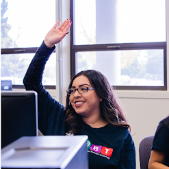 Woman in CABT t-shirt at computer raises hand and smiles