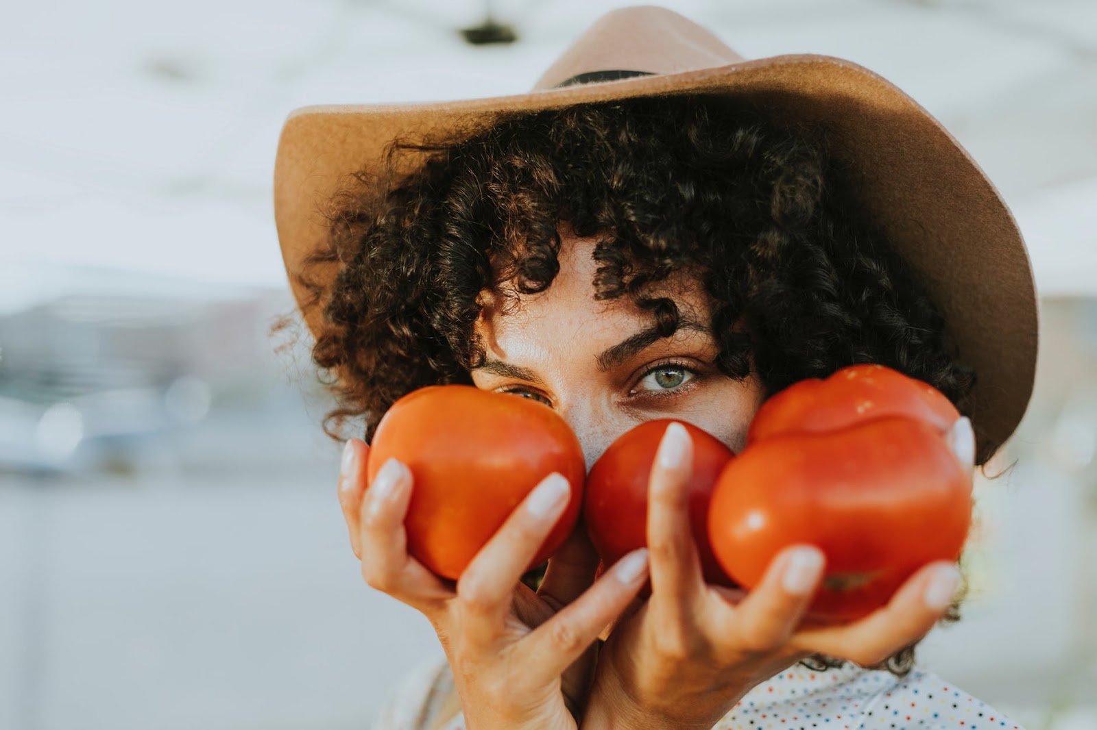 A curly haired woman with a wide hat holding tomatoes in front of her face