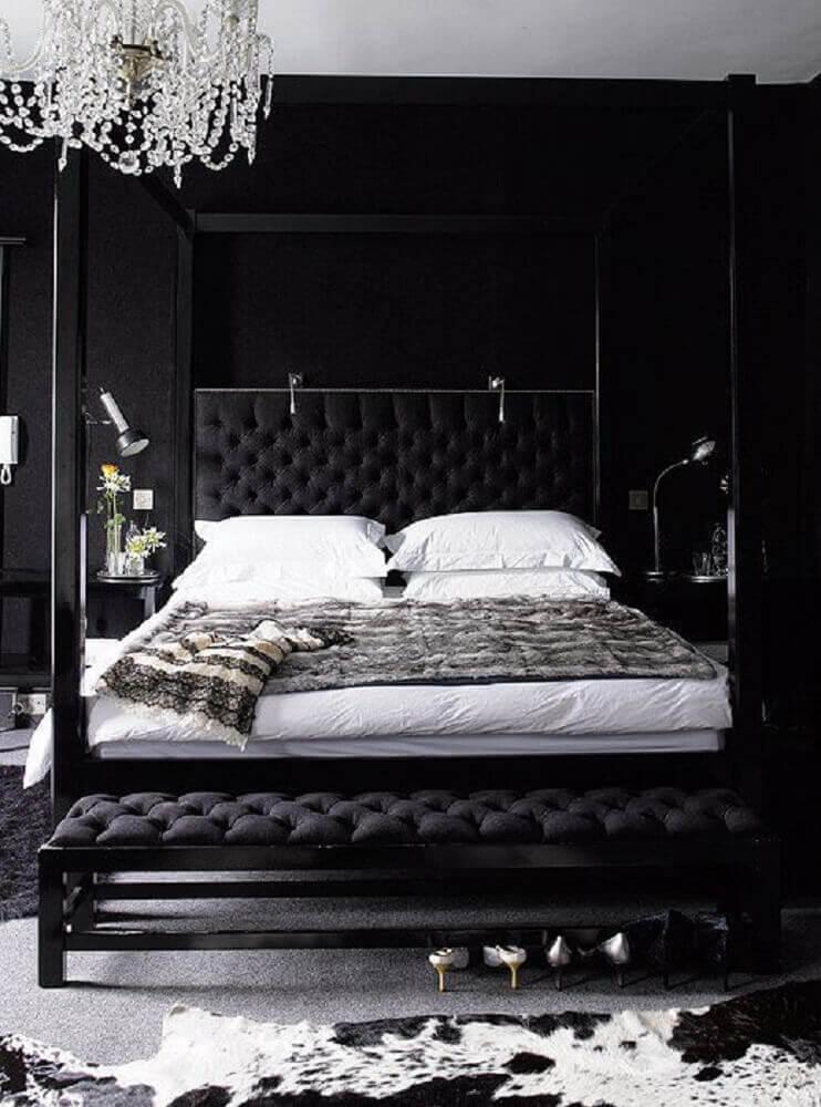 Quarto com cama de casal com cabeceira estofada preta, paredes pretas, lustre de cristal, puff no pé da cama preto e tapetes cinza.