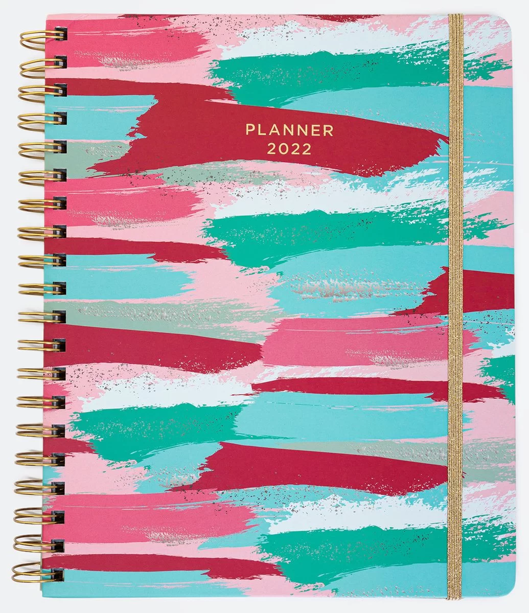 #DescriçãoDaImagem: caderno com estampa azul, rosa e verde e elástico dourado. Foto: Renner.