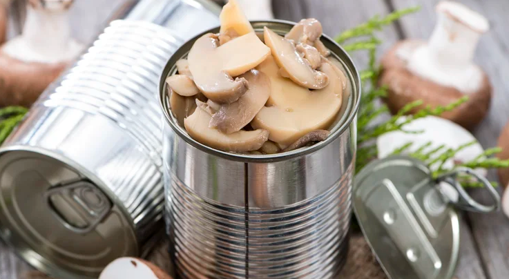 An open can of canned mushrooms, with some cans and mushrooms lying around the side of it