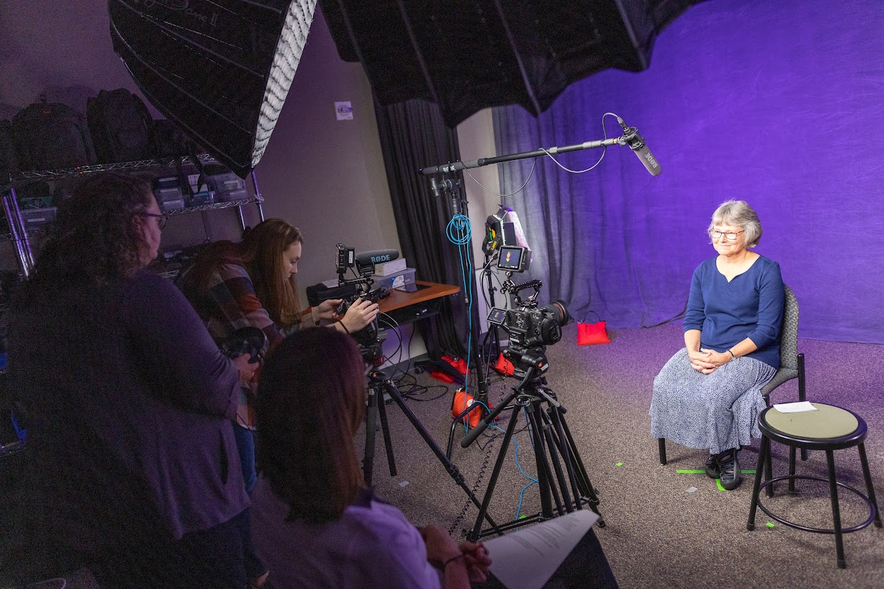 faculty member with gray hair and a gray skirt sitting in the production studio in front of a purple backdrop with three women operating video production equipment in the foreground