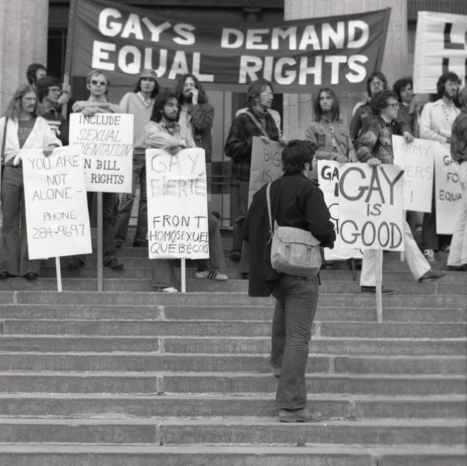Black and white photograph of the 1987 pride march, with protestors standing on the steps of the Winnipeg Legislative Building holding signs that read “gays demand equal rights” and “gay is good,” among other phrases.