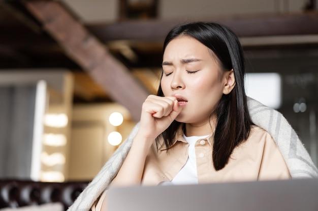 Asian woman being ill coughing at fist feeling sick at home resting with laptop