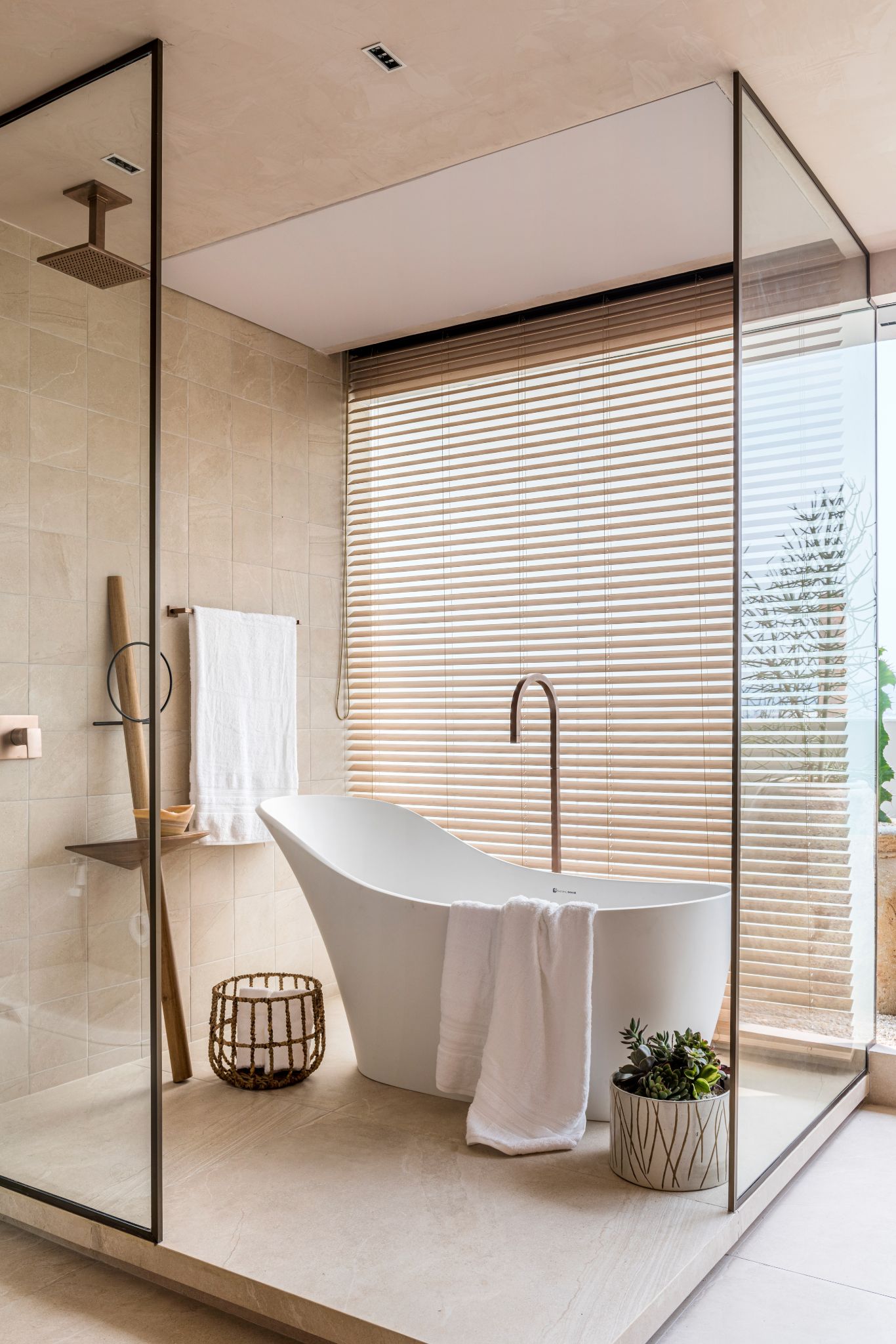 Box de vidro de banheiro com banheira de imersão branca, piso e revestimento da parede em tom nude e vasos de planta no chão