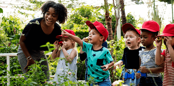 Adult woman with short dark curly hair a black shirt and grey pants with six young children wearing red caps surrounded by plants 