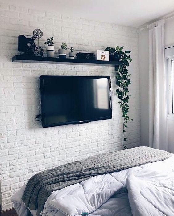 Em estilo bem escandinavo, a parede em frente a cama é de tijolinhos aparentes na cor branco, com prateleira preta, objetos, plantas e televisão.