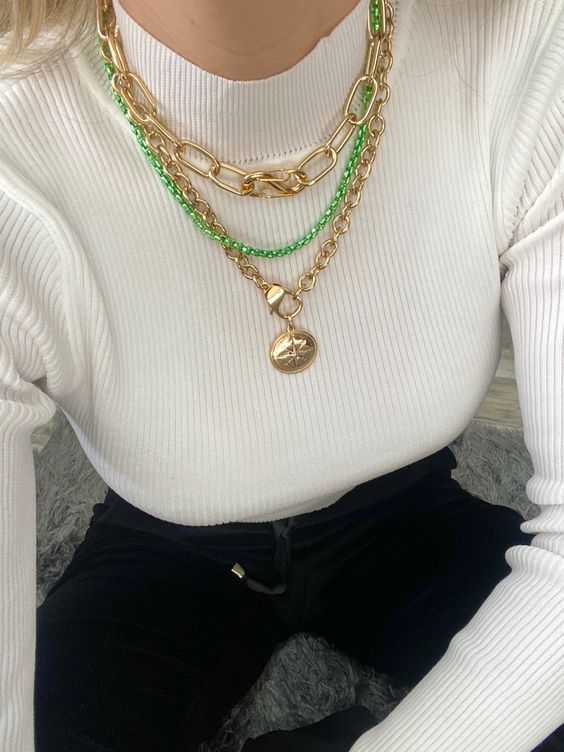 #DescriçãoDaImagem: foco da foto no corpo de uma mulher sentada que veste blusa branca de manga comprida e calça escura e tem colares em forma de corrente dourada e verde. Foto: Pinterest.