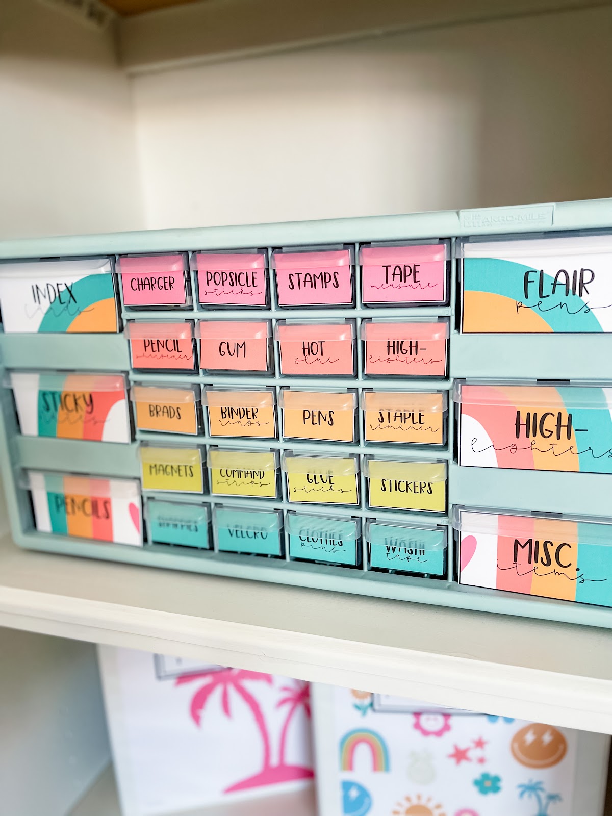 This image shows a teacher toolbox used to hold small supplies. Each drawer is labeled to display what goes in each drawer. The larger drawers on the outside are decorated with two sides of a rainbow. The boxes on the inside are simply colorful squares, no extra decorations. 