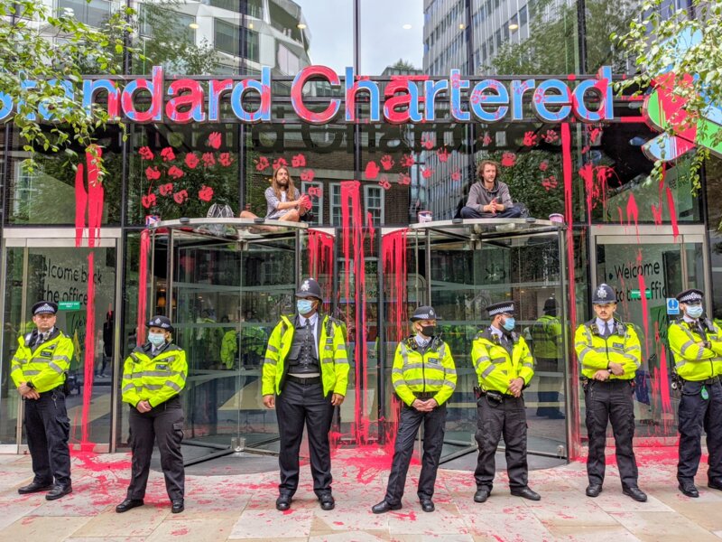 A line of police pffocers in front of the Standard Chartered building covered in fake blood, with two rebels seating on top of the entrance doors