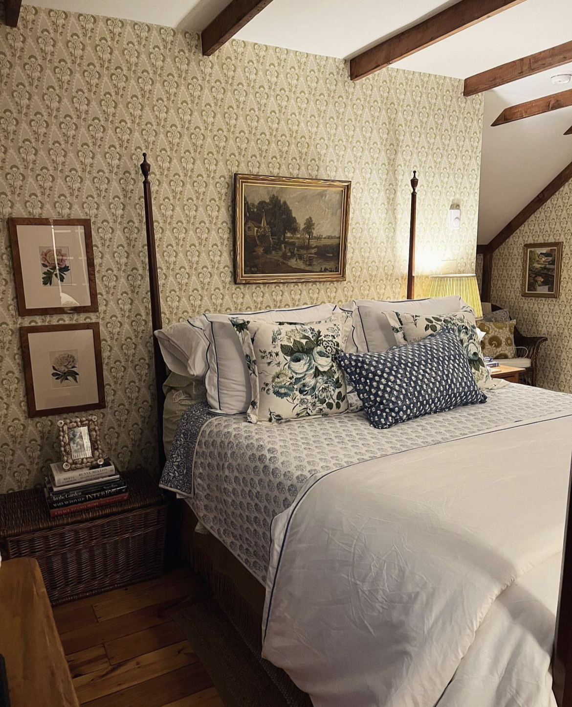 Four poster bed layered with a variety of pillows and vintage bed linens in a room with several works of art and a chest standing as a night stand.