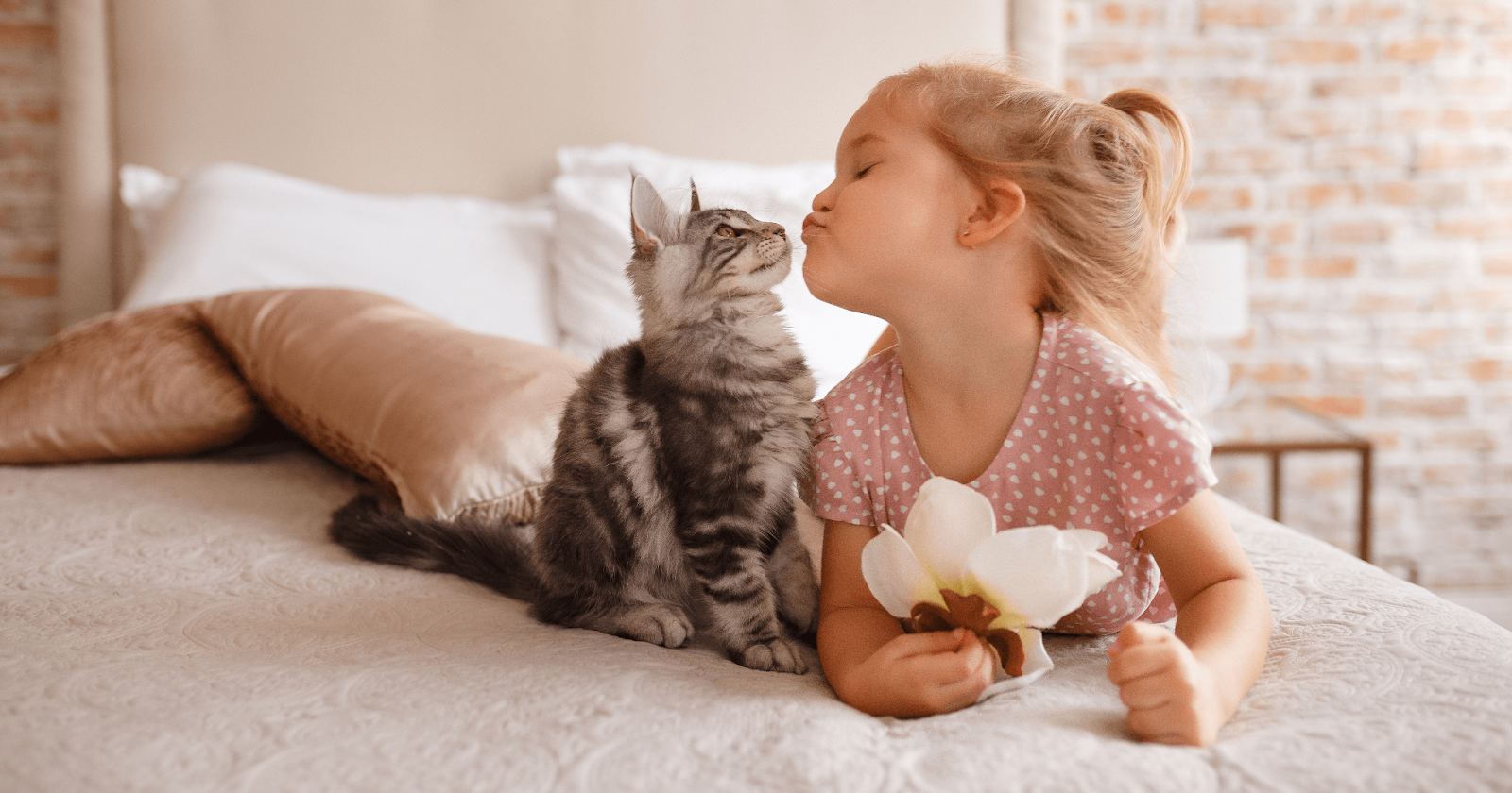 Young girl laying on a bed making a kiss face toward cat sitting beside her