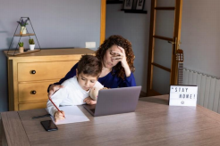Image Description: A photo of a parent and their child working at a table together. The parent is covering their face with their hand, while the child is writing on a sheet of paper next to a laptop.