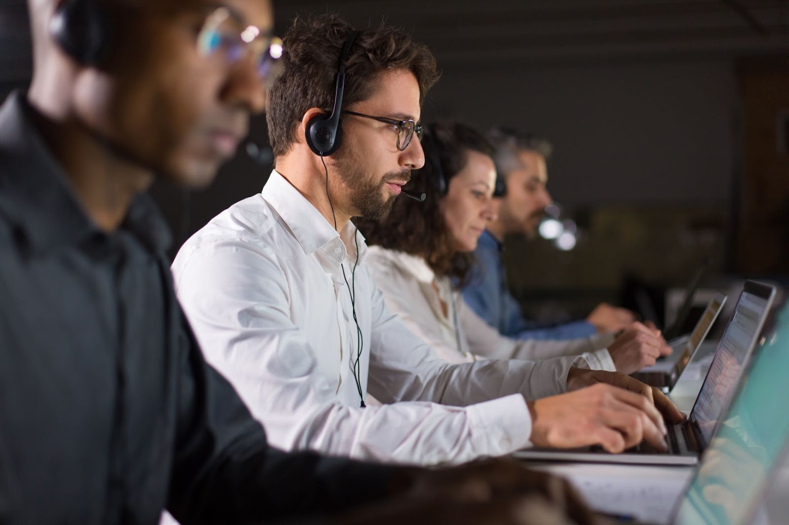 imagem com vista lateral de 4 pessoas trabalhando ao em seus notebooks. O primeiro homem é negro e usa óculos, o segundo homem é branco, com barba e óculos, em seguida temos uma mulher de cabelos enrolados e, por último, um homem de meia idade, cabelos grisalhos e barba.