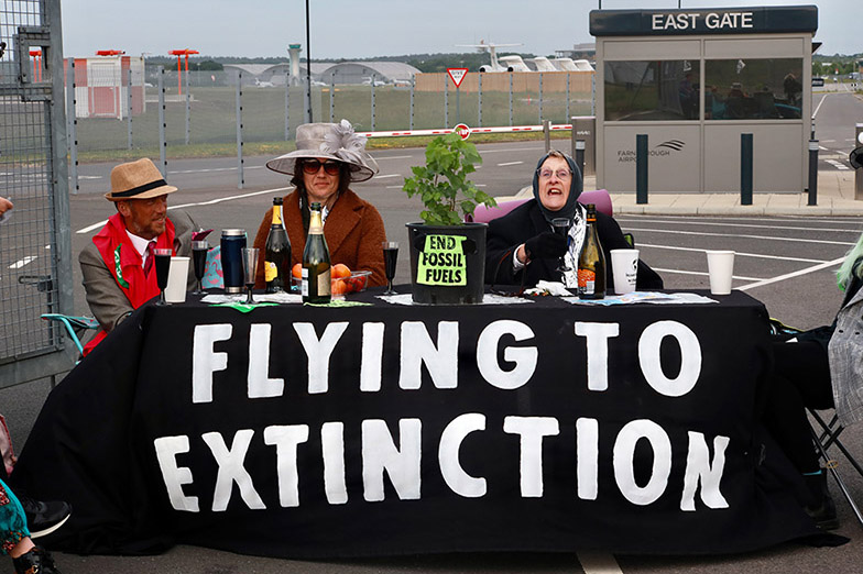 Blocking airport entrance, three rebels in fancy dress sit at a table with food and bottles and large banner that says FLYING TO EXTINCTION