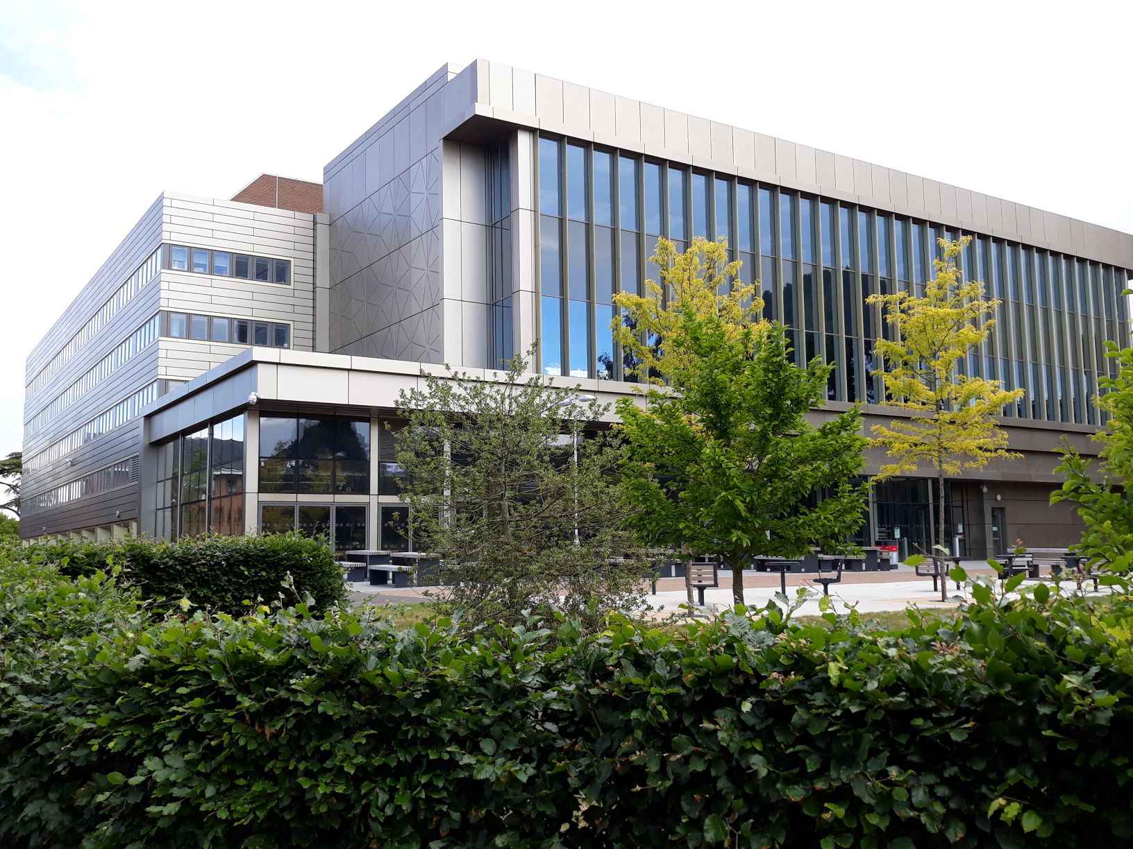 Image of the University Library. It is a large, modern building, recently refurbished. It is grey, with many windows. In front of it is green space with trees.