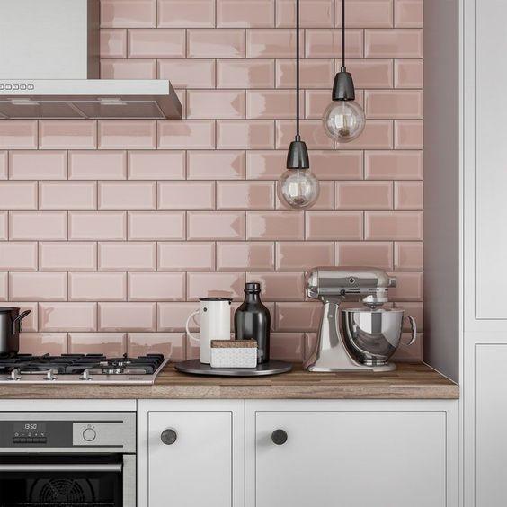 cozinha com armários brancos, bancada da pia amadeirada com eletrodomésticos de inox, luminária pendente cinza e azulejo subway tiles rosa na parede da pia