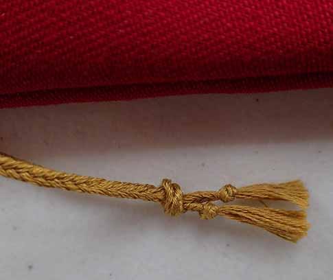 The two gold drawstrings knotted together in one larger knot above the small knots preventing fraying at the bottom of each drawstring. Side edge of the red bag is visible.