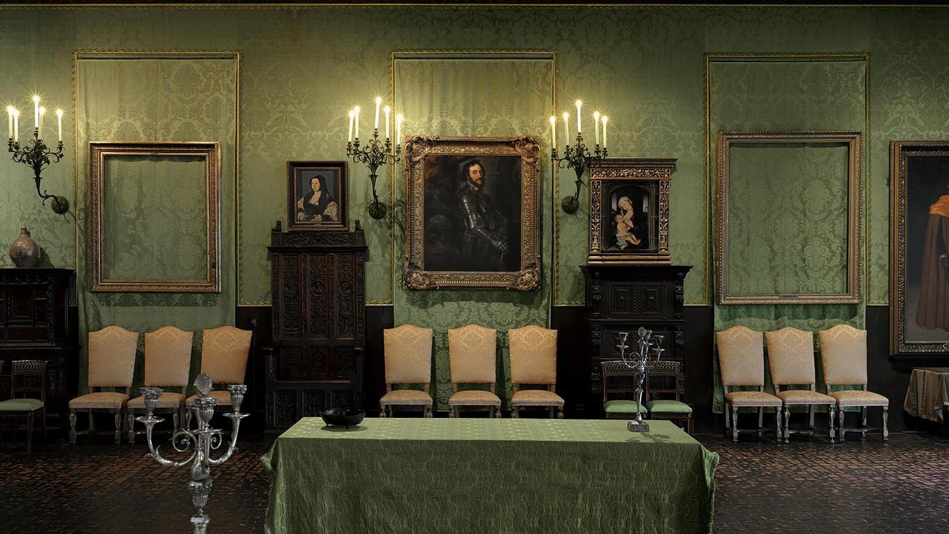 In an elegant room with green brocade wallpaper, six ornate frames are visible. The center three and the one on the far right all carry portraits, but the remaining two are empty. Decorative chairs, tables, and furniture fill the room, which is lit by candelabras. 