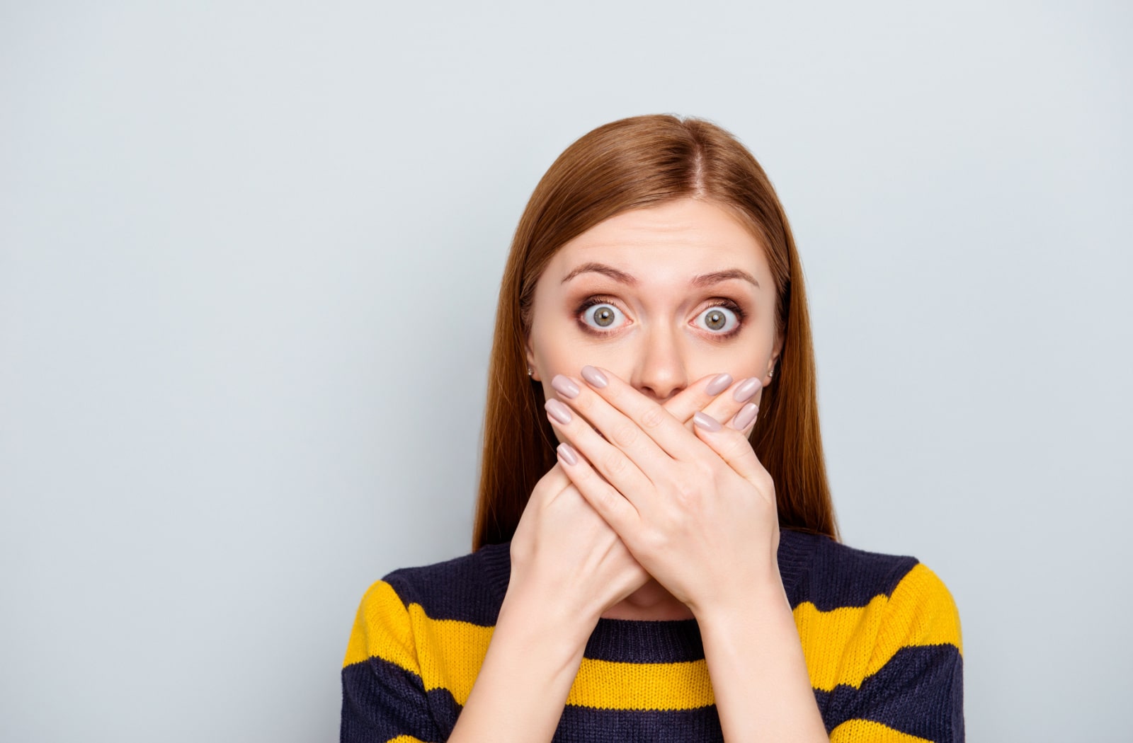 A woman wearing a black and yellow striped sweater covers her mouth with both hands, because she is surprised her breath smells unpleasant.