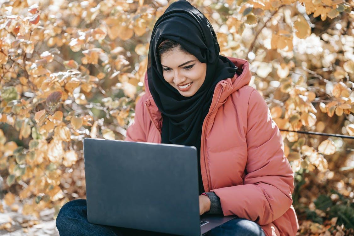 Free Cheerful young Middle East female in casual clothes and traditional headscarf browsing laptop in autumn park Stock Photo